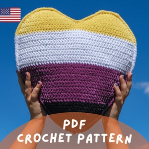 May include: A crochet heart-shaped pillow in yellow, white, purple, and black colors. The pillow is being held up by a person's hands. The text "PDF CROCHET PATTERN" is at the bottom of the image.
