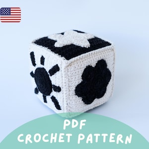 May include: A crocheted cube with black and white patterns. The cube features a star, sun, and cloud design. The text "PDF CROCHET PATTERN" is displayed on a teal background.