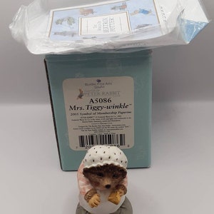 Puede incluir: Una figura de Mrs. Tiggy-winkle, un personaje de erizo de los cuentos de Beatrix Potter, se muestra. La figura es marrón, blanca y rosa, y está posada sobre una base gris. La caja tiene el texto "Mrs. Tiggy-winkle" y "Peter Rabbit Club".