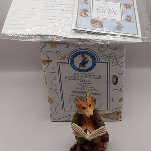 Könnte beinhalten: Eine Figur eines Fuchses, der ein Buch liest, aus der Sammlung "The World of Beatrix Potter". Der Fuchs trägt eine grüne Jacke und sitzt auf einem braunen Sockel. Die Figur steht vor einer Schachtel mit dem gleichen Thema.