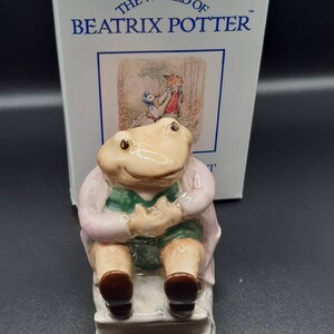 Op de afbeelding: Een keramieken beeldje van een kikker met een roze jas en groene korte broek, zittend op een kleine bank. Het beeldje komt uit de collectie "The World of Beatrix Potter", met de doos zichtbaar op de achtergrond.