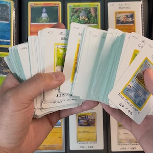 Pu&ograve; includere: Una collezione di carte collezionabili tenute in mano, con un raccoglitore sullo sfondo. Le carte sono bianche con illustrazioni colorate e testo. Alcune carte sono etichettate "Neo Discovery" e "Paldea Evolved".