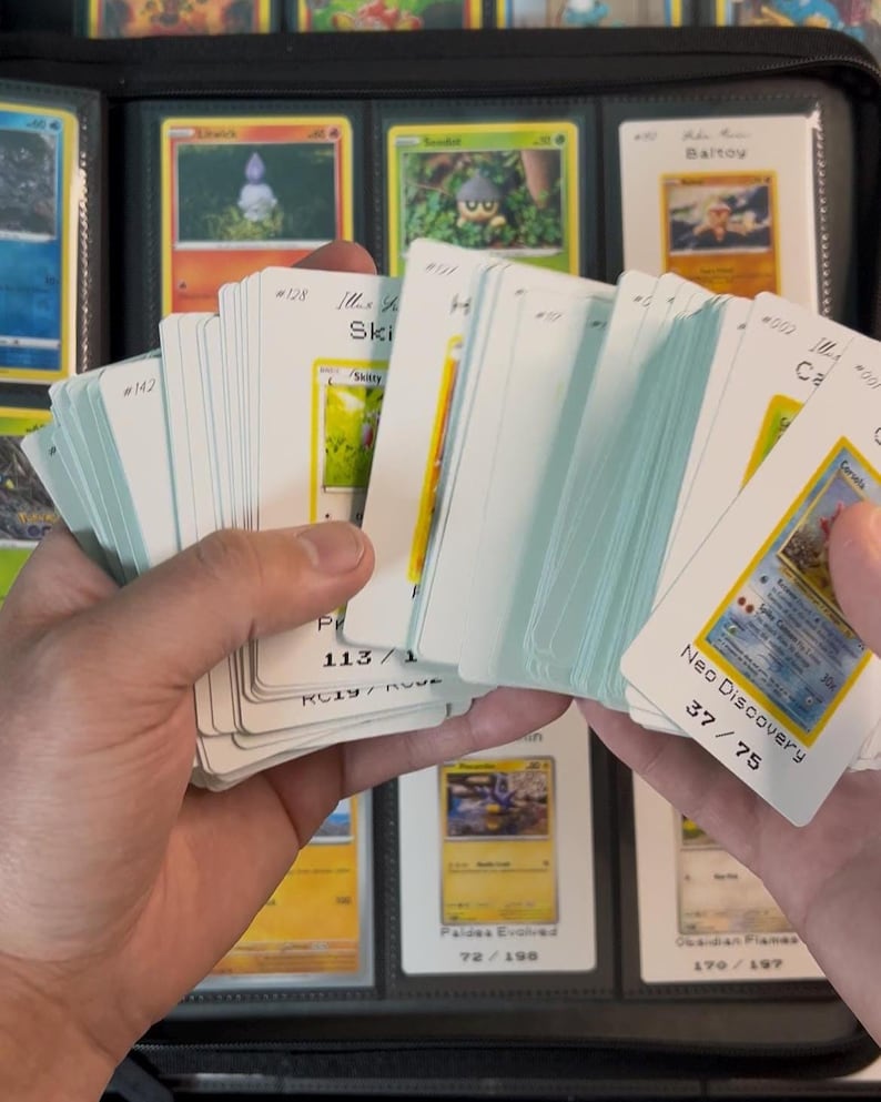 Pu&ograve; includere: Una collezione di carte collezionabili tenute in mano, con un raccoglitore sullo sfondo. Le carte sono bianche con illustrazioni colorate e testo. Alcune carte sono etichettate "Neo Discovery" e "Paldea Evolved".
