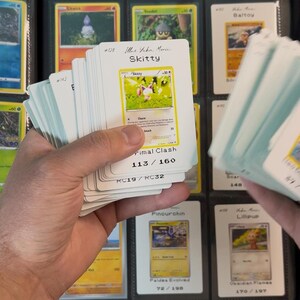 Pu&ograve; includere: Una mano che tiene in mano una pila di carte collezionabili Pok&eacute;mon, con un raccoglitore sullo sfondo. Le carte presentano vari personaggi Pok&eacute;mon e informazioni di gioco. La carta Skitty &egrave; visibile, con il testo "Skitty" e "Primal Clash".