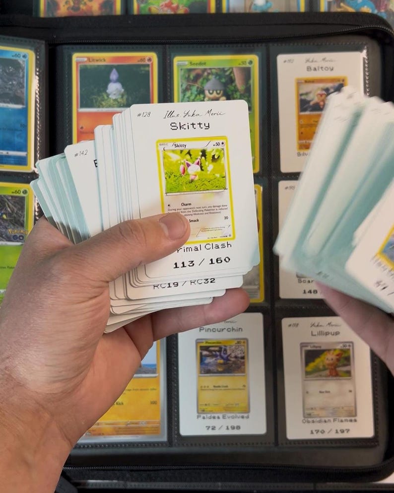 Pu&ograve; includere: Una mano che tiene in mano una pila di carte collezionabili Pok&eacute;mon, con un raccoglitore sullo sfondo. Le carte presentano vari personaggi Pok&eacute;mon e informazioni di gioco. La carta Skitty &egrave; visibile, con il testo "Skitty" e "Primal Clash".