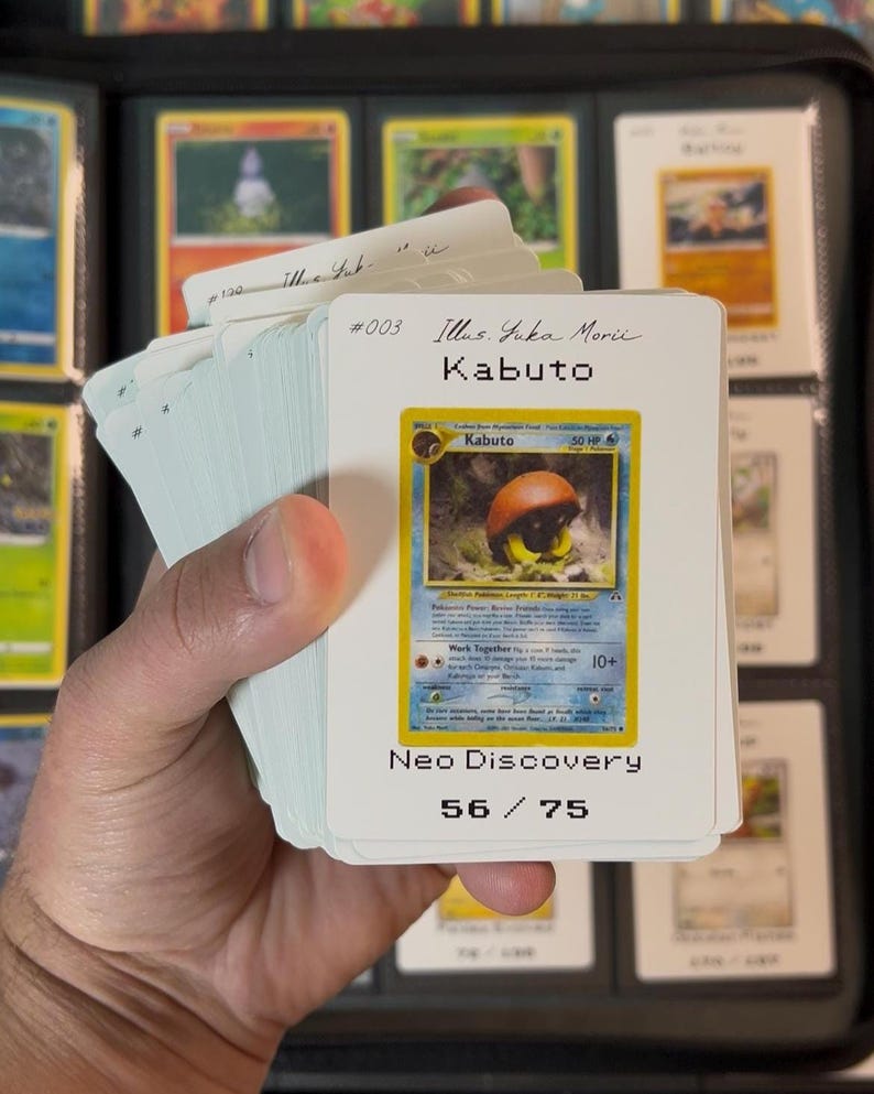 Pu&ograve; includere: Un mucchio di carte collezionabili Pok&eacute;mon, con una carta Kabuto della serie Neo Discovery. La carta mostra il nome "Kabuto" e "50 HP". Il numero del set &egrave; 56/75.
