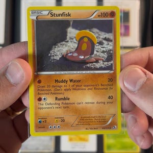 Pu&ograve; includere: Una carta collezionabile Pok&eacute;mon Stunfisk, tenuta tra due mani. La carta ha un bordo giallo e presenta un'illustrazione a cartoni animati di Stunfisk. Il testo della carta include gli attacchi "Muddy Water" e "Rumble".