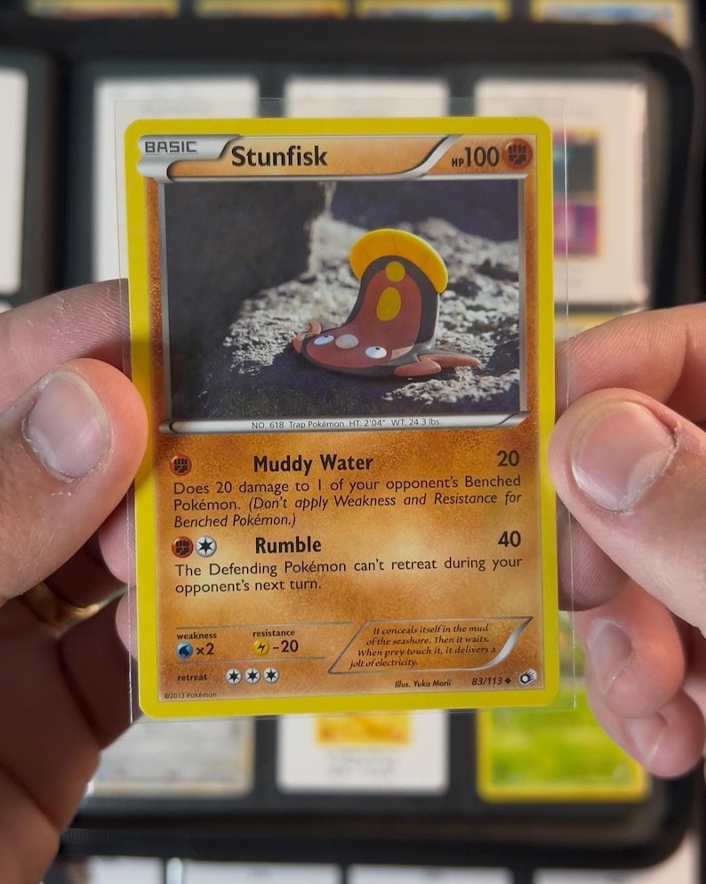 Pu&ograve; includere: Una carta collezionabile Pok&eacute;mon Stunfisk, tenuta tra due mani. La carta ha un bordo giallo e presenta un'illustrazione a cartoni animati di Stunfisk. Il testo della carta include gli attacchi "Muddy Water" e "Rumble".