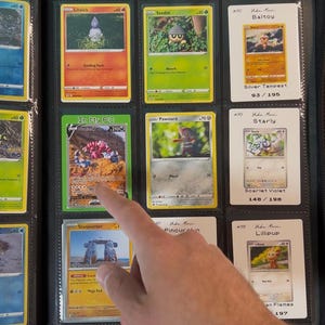 Pu&ograve; includere: Un raccoglitore nero pieno di carte collezionabili Pok&eacute;mon. Le carte sono in custodie di plastica trasparente. Le carte presentano illustrazioni colorate e testo, inclusi nomi di carte e informazioni sul set. Un dito indica una carta.