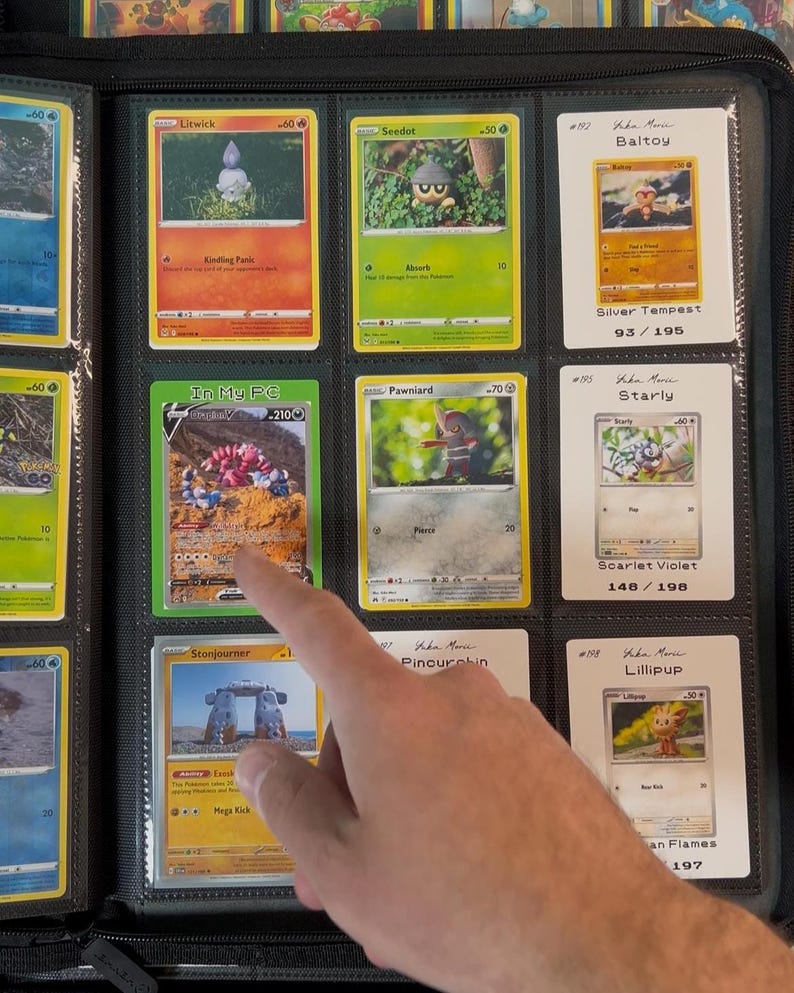 Pu&ograve; includere: Un raccoglitore nero pieno di carte collezionabili Pok&eacute;mon. Le carte sono in custodie di plastica trasparente. Le carte presentano illustrazioni colorate e testo, inclusi nomi di carte e informazioni sul set. Un dito indica una carta.