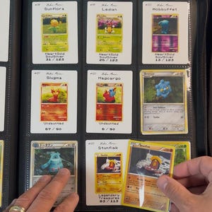 Pu&ograve; includere: Una collezione di carte collezionabili Pok&eacute;mon esposta in un raccoglitore nero. Le carte presentano vari personaggi come Sunflora, Ledian e Stunfisk, con testo che indica nomi di set e numeri di carte. Il raccoglitore &egrave; aperto e una mano tiene in mano una carta.