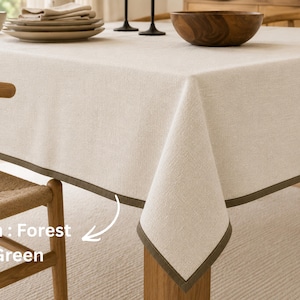May include: A rectangular beige tablecloth with a textured fabric finish and a solid forest green border trim. The tablecloth is draped over a table, featuring a clean hemmed edge that highlights the contrast between the light neutral fabric and the dark green accent colour.