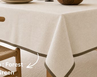 May include: A rectangular beige tablecloth with a textured fabric finish and a solid forest green border trim. The tablecloth is draped over a table, featuring a clean hemmed edge that highlights the contrast between the light neutral fabric and the dark green accent color.