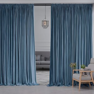 May include: A pair of floor-length, solid blue velvet curtains hanging from a black rod. The fabric features vertical pleats and a soft, light-reflecting texture. The curtains are parted in the center, revealing a glimpse of a room interior.