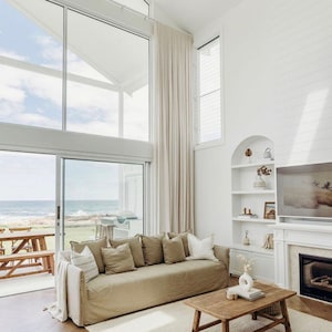 Peut inclure: Un canapé en lin beige clair avec plusieurs coussins est placé dans un salon avec une grande fenêtre donnant sur l'océan. Une table basse en bois avec un vase de fleurs séchées se trouve devant le canapé. La pièce a des murs blancs et un tapis blanc.