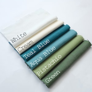 Pu&ograve; includere: Una pila di sei campioni di tessuto rettangolari con bordi sfilacciati, disposti in un motivo diagonale a cascata. I colori sono etichettati come White, Cream, Teal Blue, Aqua Blue, Pistachio e Green, ognuno con una trama tessuta visibile.