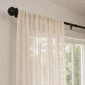 Può includere: Un pannello per tenda semitrasparente color bianco sporco con una sottile trama a righe verticali, appeso a un bastone per tende in metallo nero opaco con un terminale rotondo. Il tessuto leggero lascia passare la luce, creando un effetto soffuso.