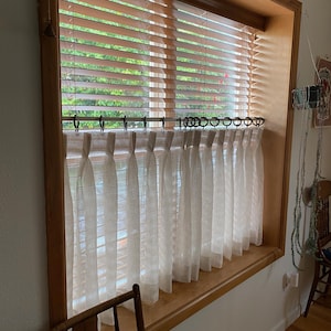 Pode incluir: Cortinas brancas transparentes com um topo franzido e uma moldura de janela de madeira com persianas.