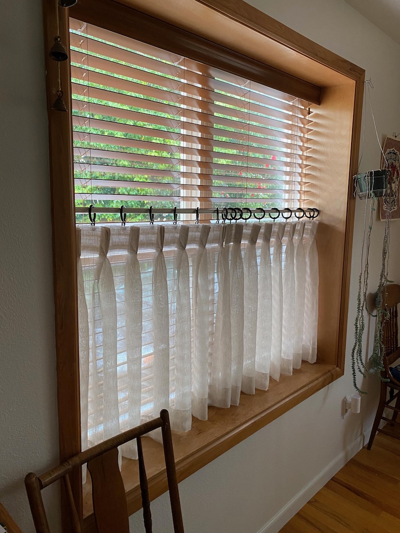 Pode incluir: Cortinas brancas transparentes com um topo franzido e uma moldura de janela de madeira com persianas.