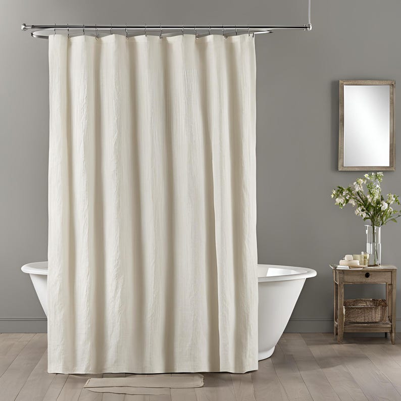 Pu&ograve; includere: Una tenda da doccia color bianco sporco con una sottile trama verticale stropicciata, appesa a un'asta in metallo cromato lucido tramite anelli argentati. Il tessuto cade in pieghe uniformi, creando un aspetto pulito e semplice.