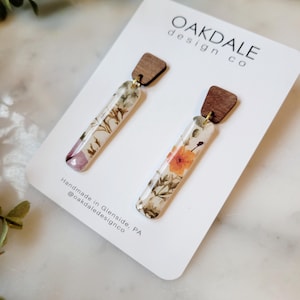 May include: A pair of long, rectangular earrings with pressed flowers encased in clear resin. The earrings have a wood top and are handmade in Glenside, PA. The text "Oakdale design co" is printed on the white card.