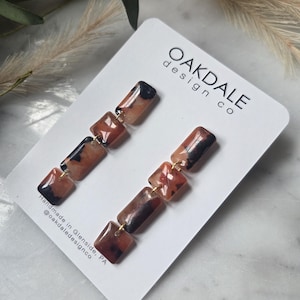 Op de afbeelding: Hangende oorbellen met een rechthoekig ontwerp in bruin, zwart en beige. De oorbellen zijn verbonden door goudkleurig metaal. Ze worden gepresenteerd op een witte kaart met de tekst "OAKDALE design co".