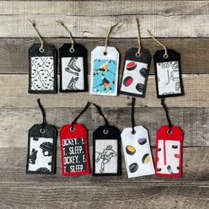 May include: Ten felt gift tags with hockey-themed designs. The tags are black, white, red, and blue with images of hockey players, pucks, and skates. Some tags have text that says "Hockey, Eat, Sleep, Hockey, Repeat".