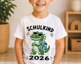 Pode incluir: Uma t-shirt branca de manga curta com um dinossauro verde de desenho animado a usar óculos de sol pretos e uma mochila azul. O texto SCHULKIND está impresso a preto acima do dinossauro e o número 2026 está impresso a preto abaixo.