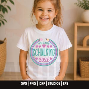 May include: A white t-shirt with a colorful graphic design featuring the text "ERSTKLASSIG SCHULKIND 2026" in a circular design with hearts and a pencil illustration. The shirt is worn by a young child.