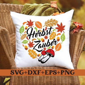 Puede incluir: Cojín cuadrado blanco con un diseño en forma de corazón de hojas de otoño, bellotas y setas. Las palabras "Herbst Zauber" están en el centro. La parte inferior del cojín tiene el texto "SVG+DXF+EPS+PNG".