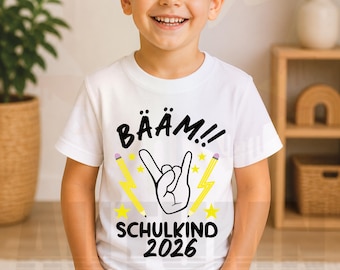 May include: A white t-shirt with a graphic design featuring the word "BÄÄM!!" above a hand gesture, pencils, lightning bolts, and stars. The text "SCHULKIND 2026" is below the graphic. The shirt is worn by a smiling child.