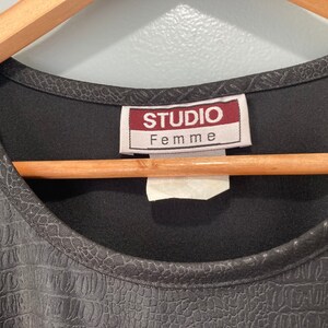Puede incluir: Camiseta negra con un patr&oacute;n texturizado similar al de cocodrilo. La camiseta tiene un escote redondo y una etiqueta que dice "Studio Femme".