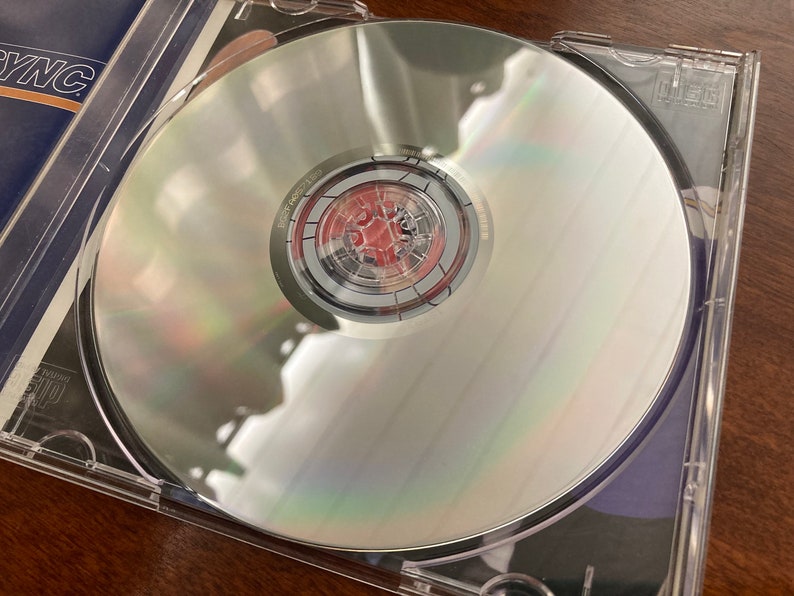 K&ouml;nnte beinhalten: Eine silberne CD in einem durchsichtigen Plastik-Etui. Die CD hat eine silberne Mitte mit einem roten und schwarzen Design.