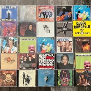 K&ouml;nnte beinhalten: Eine Sammlung von 18 Compact Discs (CDs) mit verschiedenen K&uuml;nstlern und Albumtiteln. Die CDs sind in einem Raster auf einer Holzoberfl&auml;che angeordnet. Einige der K&uuml;nstler sind Will Smith, R.E.M., No Doubt und Offspring.