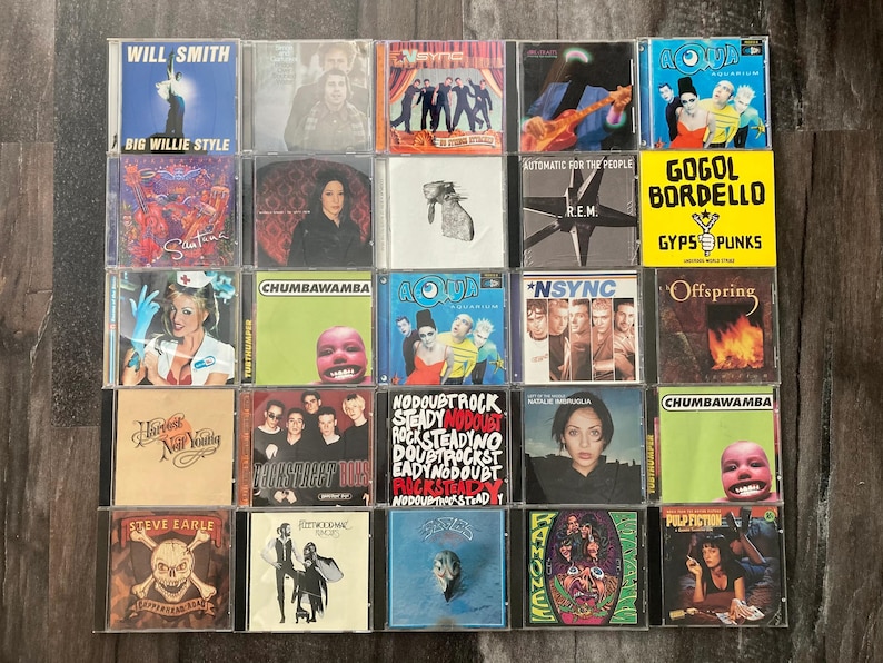 K&ouml;nnte beinhalten: Eine Sammlung von 18 Compact Discs (CDs) mit verschiedenen K&uuml;nstlern und Albumtiteln. Die CDs sind in einem Raster auf einer Holzoberfl&auml;che angeordnet. Einige der K&uuml;nstler sind Will Smith, R.E.M., No Doubt und Offspring.