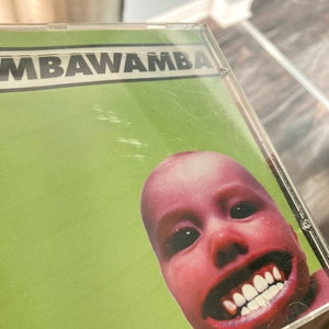 K&ouml;nnte beinhalten: Ein gr&uuml;nes CD-Geh&auml;use mit dem Titel "MBAWAMBA" in schwarzen Buchstaben. Die Vorderseite zeigt eine Nahaufnahme eines Babygesichts mit einem breiten, zahnigen Grinsen.