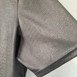 Puede incluir: Primer plano de una camisa negra de cuero sint&eacute;tico con un patr&oacute;n texturizado similar al de cocodrilo. La camisa tiene mangas cortas y un ajuste cl&aacute;sico.