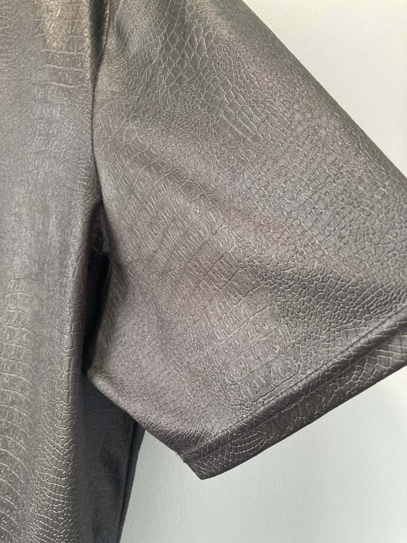 Puede incluir: Primer plano de una camisa negra de cuero sint&eacute;tico con un patr&oacute;n texturizado similar al de cocodrilo. La camisa tiene mangas cortas y un ajuste cl&aacute;sico.