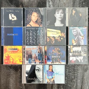 K&ouml;nnte beinhalten: Eine Sammlung von 12 Compact Discs (CDs) mit verschiedenen K&uuml;nstlern, darunter Mariah Carey, Backstreet Boys, Hanson und Monica. Die CDs sind in einem Raster auf einer Holzoberfl&auml;che angeordnet.