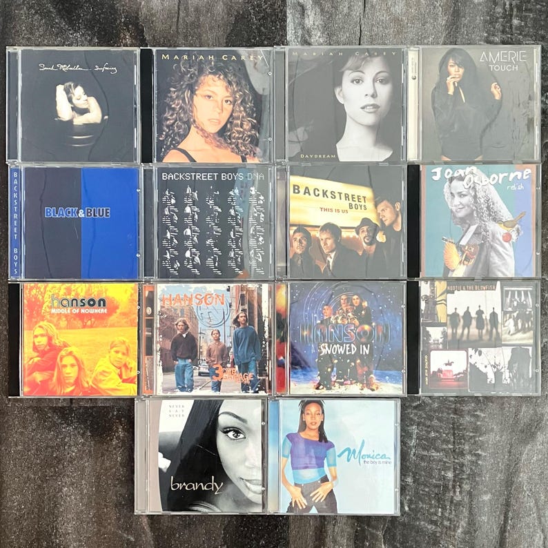 K&ouml;nnte beinhalten: Eine Sammlung von 12 Compact Discs (CDs) mit verschiedenen K&uuml;nstlern, darunter Mariah Carey, Backstreet Boys, Hanson und Monica. Die CDs sind in einem Raster auf einer Holzoberfl&auml;che angeordnet.