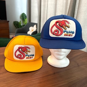 Puede incluir: Dos gorras de camionero, una amarilla y otra azul, cada una con un parche blanco con un diseño de enchufe rojo estilizado y el texto "PLUG ME!". Las gorras tienen la parte trasera de malla y viseras curvas.