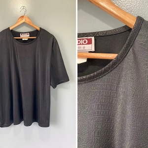 Puede incluir: Camiseta negra de manga corta con cuello redondo y etiqueta Studio One. La camiseta tiene un acabado texturizado, similar al cuero falso.