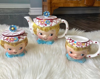 Puede incluir: Un juego de té vintage con tres piezas: una tetera, una jarra de crema y un azucarero. Cada pieza tiene forma de cabeza de niña con un sombrero blanco y rojo, ojos azules y un lazo rosa. La tetera tiene un asa roja y blanca.