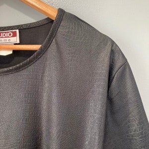 Puede incluir: Camiseta negra de manga corta con textura de piel de cocodrilo. La camiseta tiene un cuello redondo y una etiqueta que dice "Studio" con la palabra "mme" debajo.