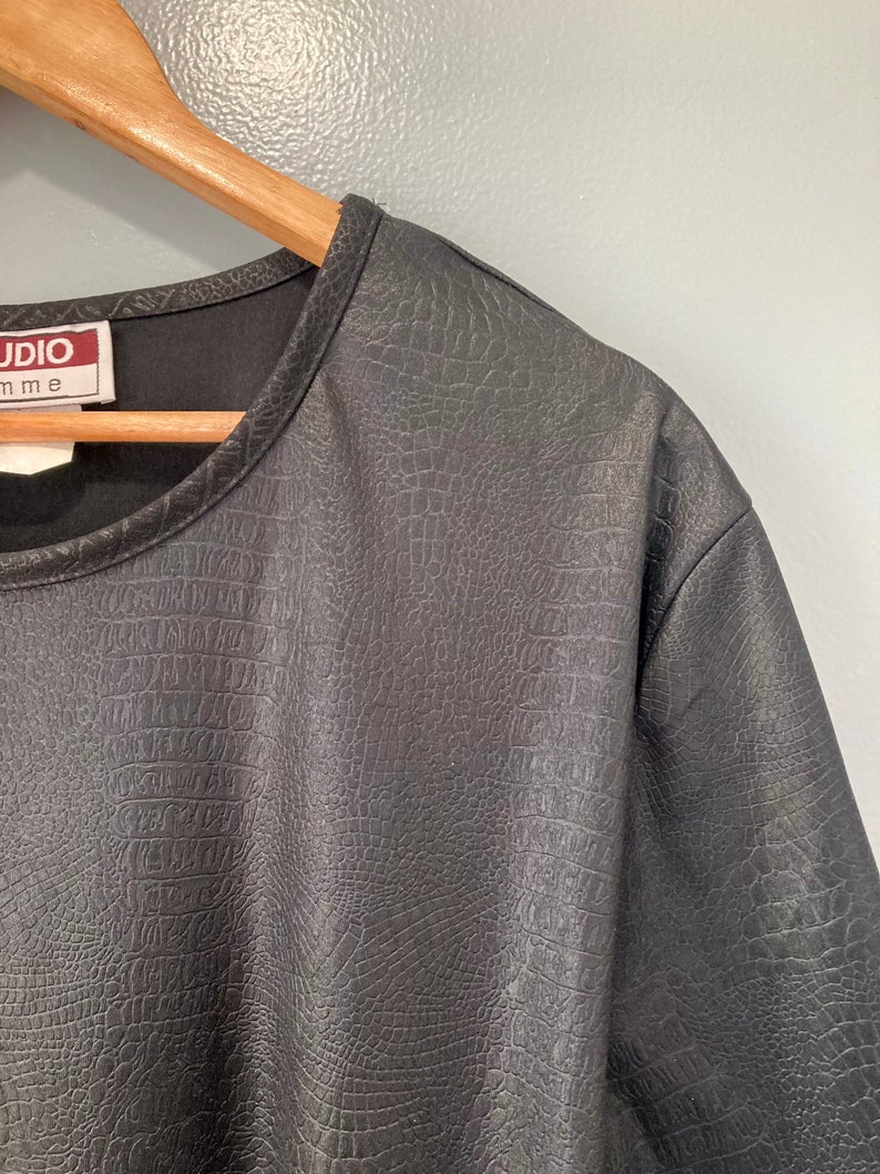 Puede incluir: Camiseta negra de manga corta con textura de piel de cocodrilo. La camiseta tiene un cuello redondo y una etiqueta que dice "Studio" con la palabra "mme" debajo.