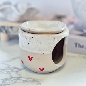 May include: A ceramic wax warmer featuring a two-tone design with a speckled white upper section and a smooth cream-colored base. The lower portion is decorated with small red heart motifs, and a large circular opening is visible on the side.