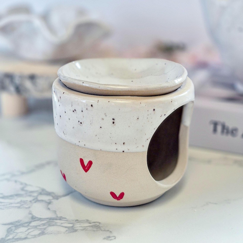May include: A ceramic wax warmer featuring a two-tone design with a speckled white upper section and a smooth cream-colored base. The lower portion is decorated with small red heart motifs, and a large circular opening is visible on the side.