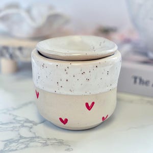 May include: A ceramic wax warmer featuring a two-tone design with a speckled white upper section and a smooth beige lower section. The lower portion is decorated with small, hand-painted red hearts. A matching speckled white lid sits on top.