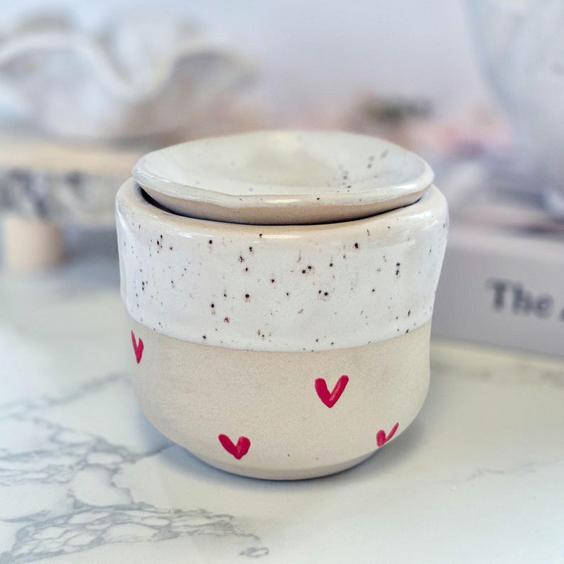 May include: A ceramic wax warmer featuring a two-tone design with a speckled white upper section and a smooth beige lower section. The lower portion is decorated with small, hand-painted red hearts. A matching speckled white lid sits on top.