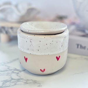 May include: A ceramic wax warmer featuring a cylindrical base with a matte beige finish decorated with small hand-painted red hearts. The upper section has a white speckled glaze and includes a matching removable ceramic lid on top.
